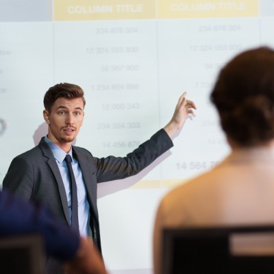 Serious middle-aged businessman standing at projection screen with table and pointing to it while looking and explaining ideas to audience seen partly